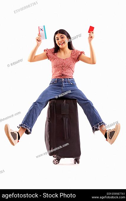 An excited woman sitting on her luggage holding credit card and air travel ticket