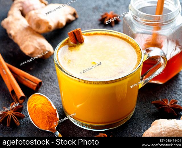 Healthy drink golden turmeric latte in glass cup.Gold milk with turmeric, ginger root, cinnamon sticks, dried turmeric powder and honey over black cement...