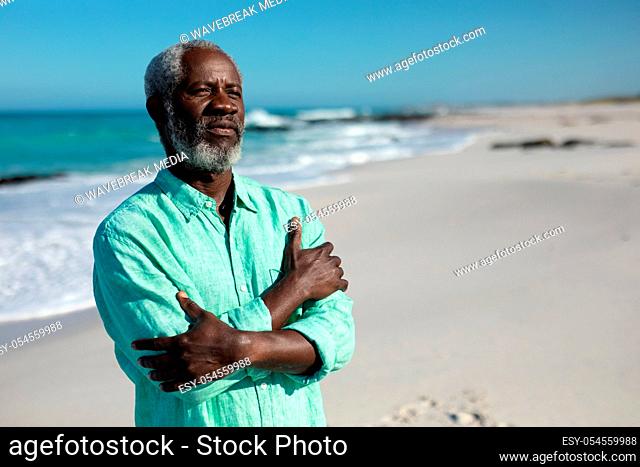 Portrait of a senior African American man standing on the beach with blue sky and sea in the background, looking into the distance with his arms crossed