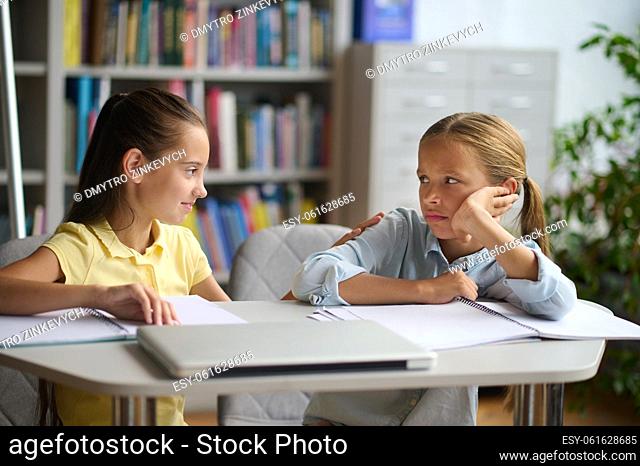 Friendly dark-haired schoolgirl sitting at the library table and tapping her upset classmate on the shoulder