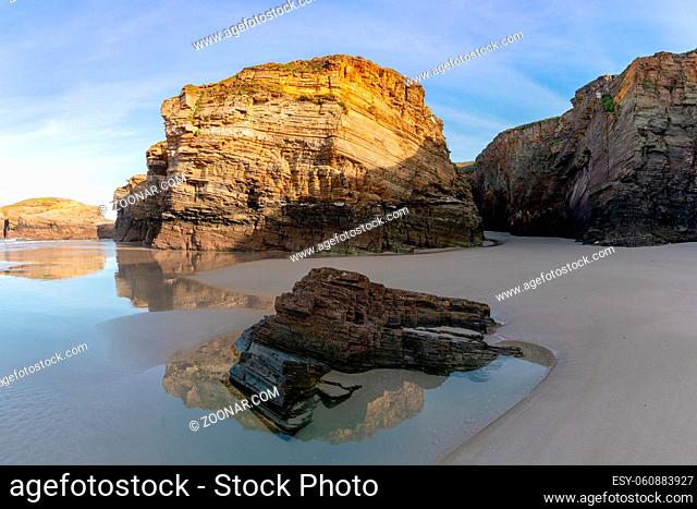 A sandy beach panorama with tidal pools and jagged broken cliffs behind in warm evening light