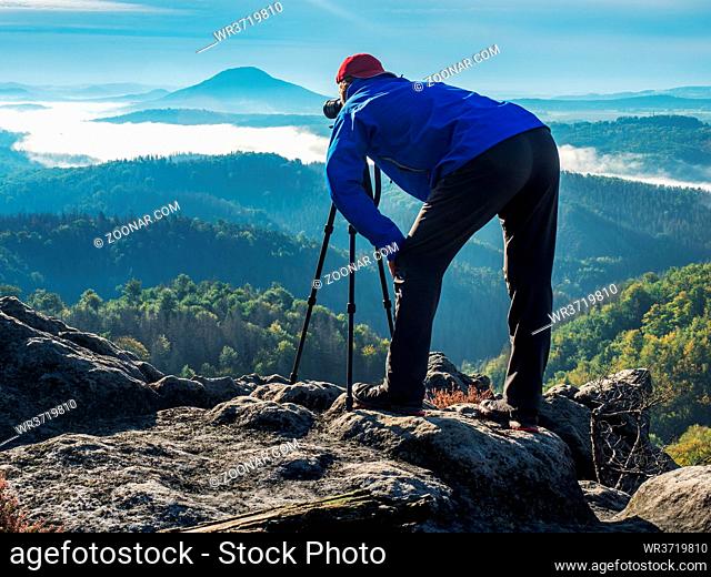 Bent photographer framing scene in viewfinder. Man is using camera on tripod for taking pictures of misty mountains scenery
