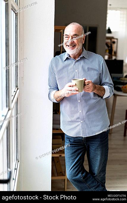 Happy businessman holding coffee mug leaning on wall at home