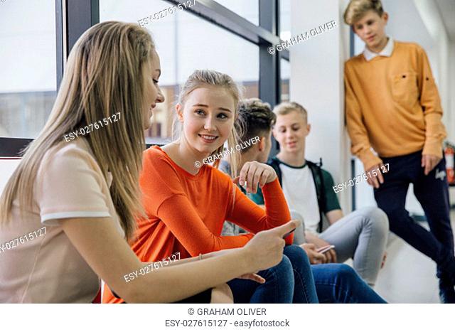 Teenagers are hanging out at school during their lunch break. Two girls are talking and looking at a smart phone together and some boys are in the back ground...