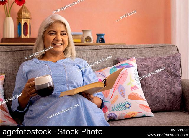 Cheerful old woman drinking tea while sitting on sofa at home