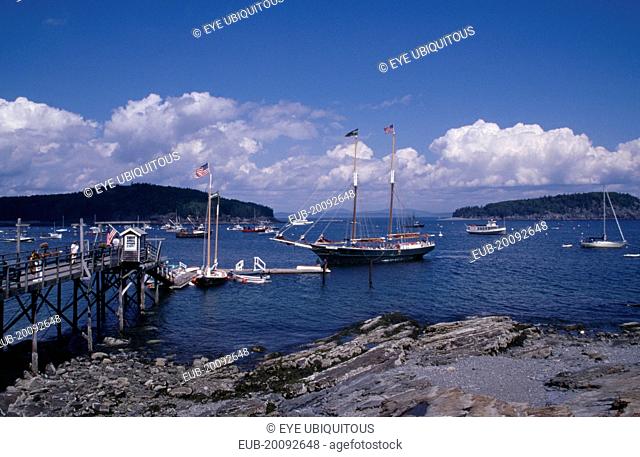 Wooden jetty with approaching yacht, various boats on water and tree covered headland beyond