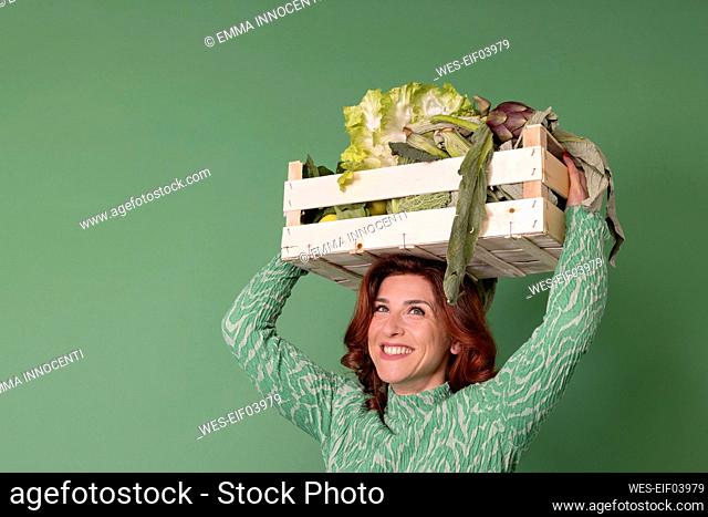 Smiling woman carrying wooden crate with vegetables against green background