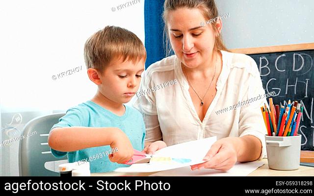 Young female teacher showing her little student how to use scissors. Boy cutting paper with scissors while doing homework