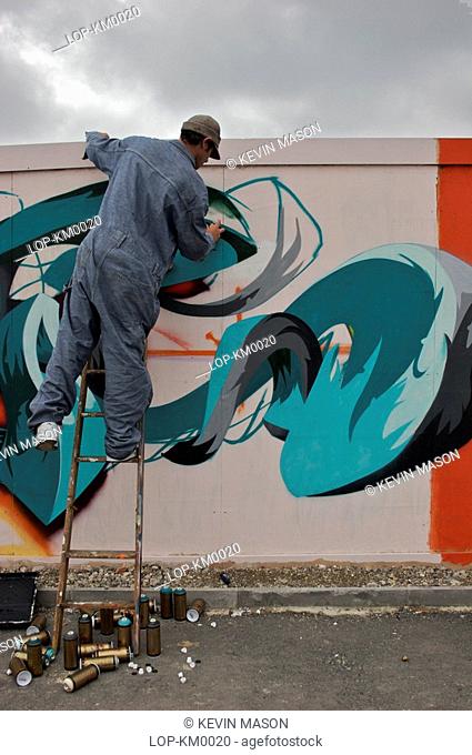 England, East Sussex, Brighton, Graffiti artist leans off a ladder to paint. The word 'graffiti' is the plural of 'graffito' and originates from the Italian...
