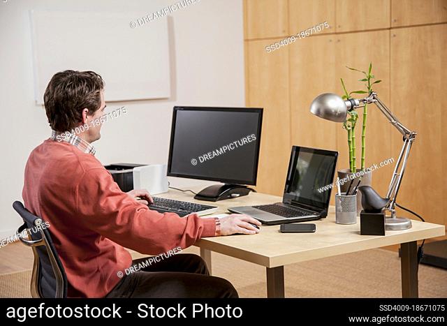 Caucasian man working on a desktop computer and laptop in home office
