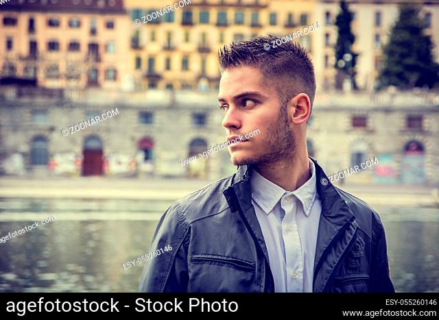 Three-quarter length of contemplative light brown haired young man wearing grey jacket and denim jeans standing beside picturesque river in Turin, Italy