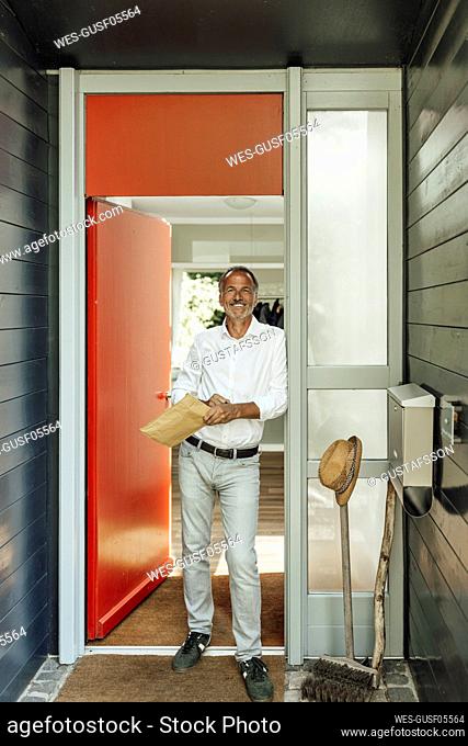 Smiling mature man holding parcel while leaning at doorway