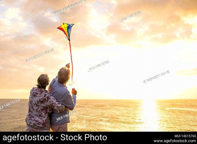 close up and portrait of two old and mature people playing and enjoying with a flaying kite at the beach with the sea at the background with sunset - active...