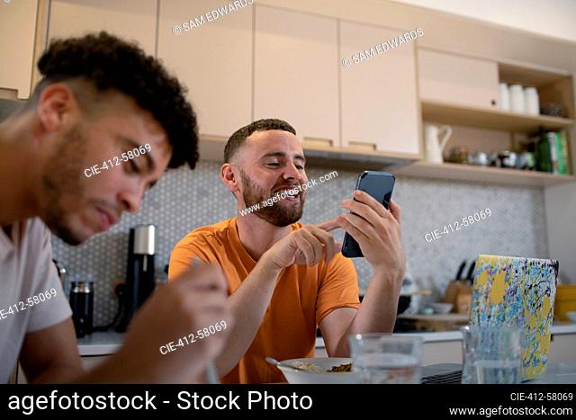 Happy gay male couple using smart phone and eating in kitchen