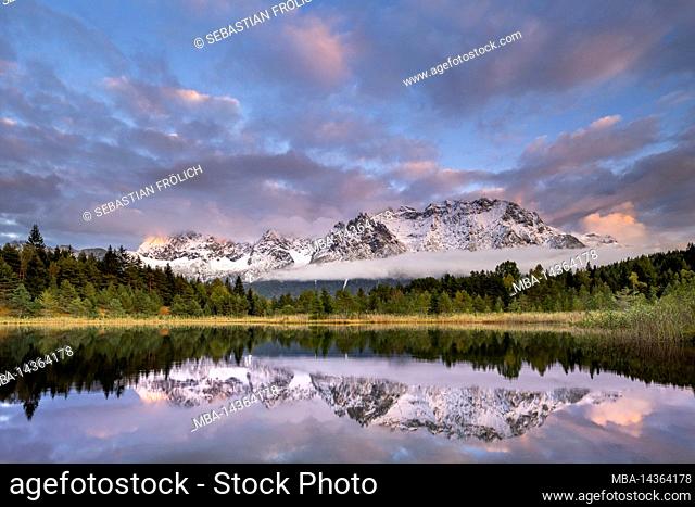 The Karwendel mountains are reflected in the Luttensee lake near Mittenwald during sunset. Fresh snow and atmospheric clouds frame the mountains
