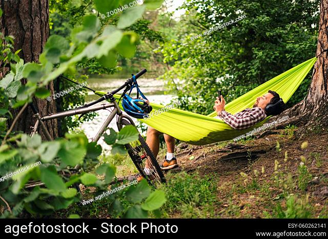 Outdoors and adventure concept. Bike trip to forest. Cyclist is resting in green hammock between trees in nature by lake while listening to music on headphones...