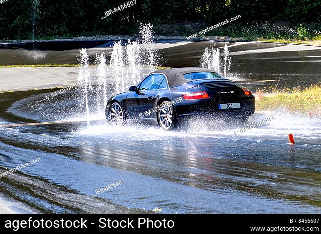 Sports car with all-wheel drive Porsche 911 Carrera 4s drives at 50 kmh speed on sloping heavily watered wet road towards obstacle from water fountain makes...