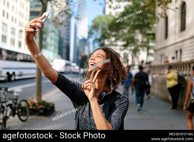 Woman showing peace sign while taking selfie through mobile phone in city