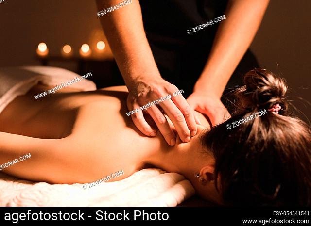 Men's hands make a therapeutic neck massage for a girl lying on a massage couch in a massage spa with dark lighting. Close-up. Dark Key