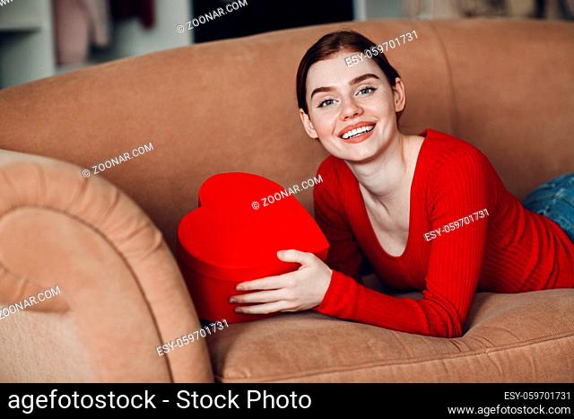 Beautifil young woman with red hair lying in her sofa in living room and holding the gift box in the shape of heart and smile. Valentines Day