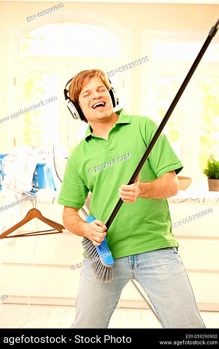 Smiling man singing with headphones, imitating playing guitar on broom at home