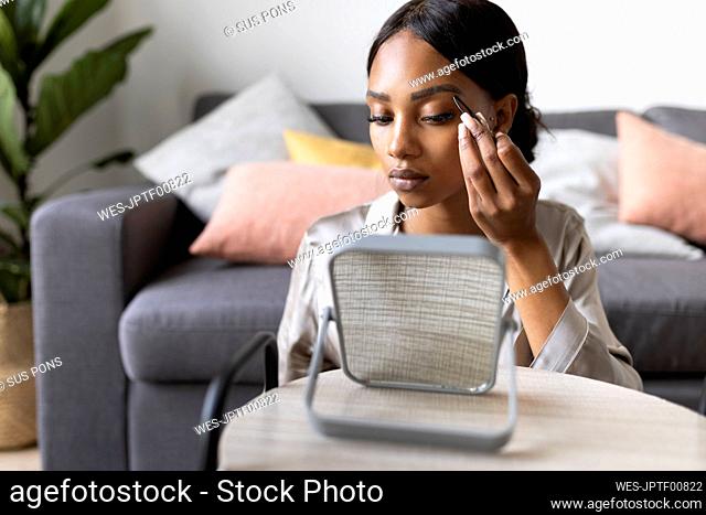 Young woman using tweezers on eyebrow while looking at mirror in living room