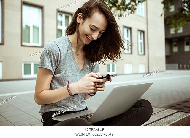 Happy young woman sitting on bench using smartphone and laptop