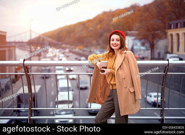 Portrait of a pretty smiling french young woman looking at camera standing at pedestrian bridge with cars on the road. Looking happy fashion girl in a ed beret...