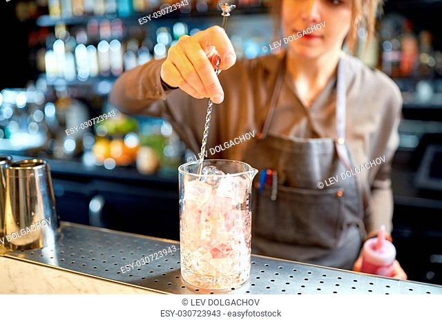 alcohol drinks, people and luxury concept - woman bartender with stirrer and glass of ice preparing cocktail at bar counter