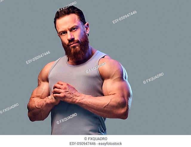Portrait of brutal bearded bodybuilder dressed in a sleeveless shirt isolated on grey background