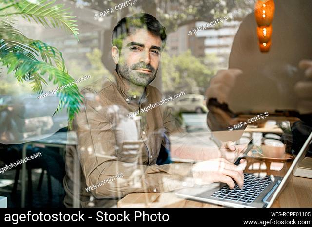 Businessman using laptop at table in restaurant seen through window glass