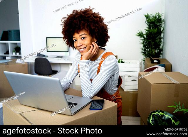 Smiling young woman with hand on chin standing by laptop in living room at new home