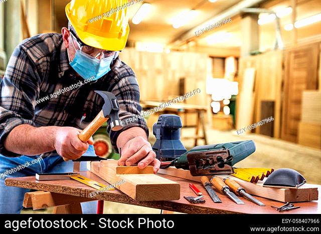 Carpenter worker at work in the carpentry workshop, wears helmet, goggles and surgical mask to prevent coronavirus infection