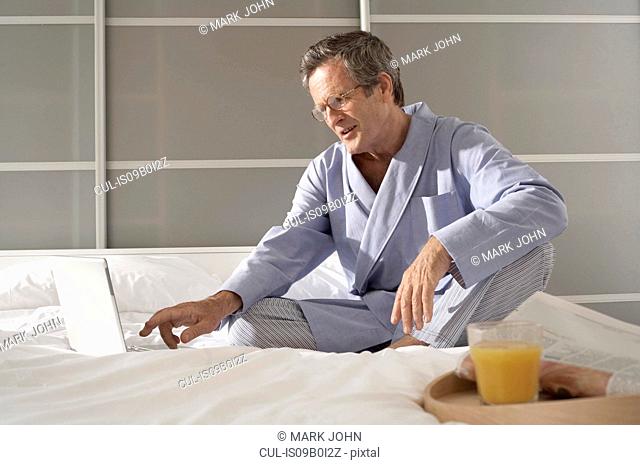 Puzzled senior man on bed typing on laptop