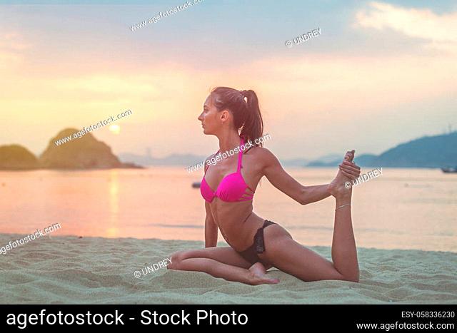 Young woman in pink bra and black panties sitting on beach stretching her legs during sunset at sea. Fitness girl doing exercises on seashore
