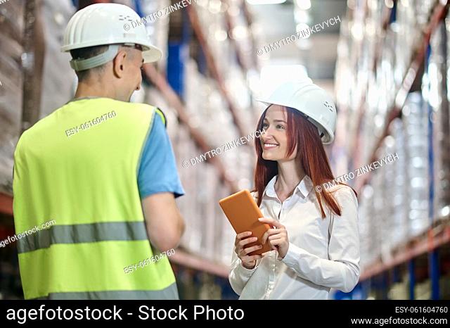 Positive communication. Smiling woman in safety helmet with tablet looking at man standing with back to camera chatting in warehouse
