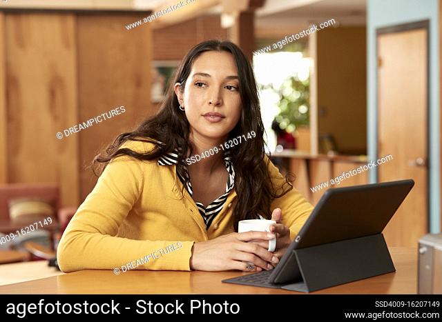 Portrait of young ethnic woman wearing yellow sweater with black and white striped blouse, sitting at bar in kitchen of downtown loft with iPad and coffee mug