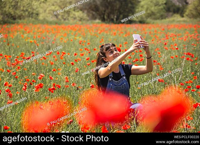 Smiling young woman taking selfie through smart phone amidst field of poppies