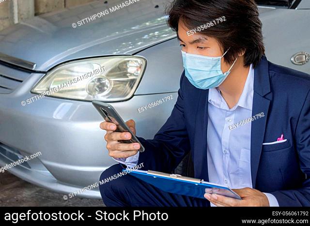 Sitting Asian Insurance Agent or Insurance Agency in Suit Wear Mask and See Smartphone and Hold Report Inspecting Car from Accident for Claim at Garage