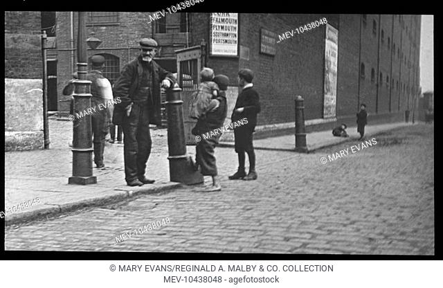 Adults and children on a London street, outside the gates of a large building, possibly a warehouse. One boy, carrying a toddler, has bare feet