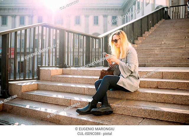 Young woman sitting on city stairway looking at smartphone, Milan, Italy