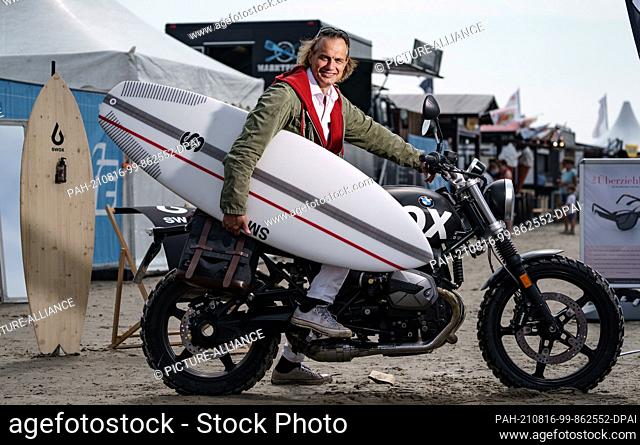 12 August 2021, Schleswig-Holstein, Sankt Peter-Ording: Actor Ralf Bauer sits on a motorbike on the beach of Sankt Peter-Ording with a surfboard under his arm