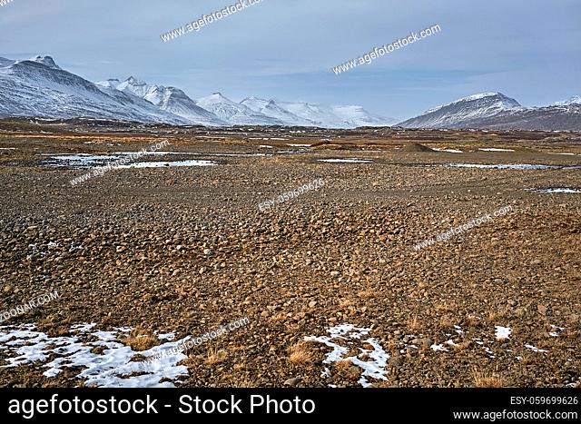 Rocky valley with remains of snow and water puddles on the background of the snowy mountains and cloudy sky in Iceland. Panoramic horizontal photo