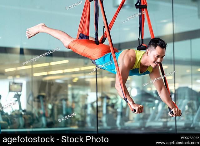 Full length low-angle view of a fit handsome man smiling while practicing anti-gravity yoga in a trendy fitness club with modern equipment