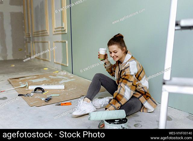 Smiling woman with paint roller having coffee in messy apartment