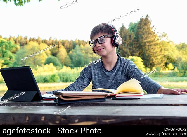 Happy male pupil in glasses, with big earphones sitting at table full of textbooks and looking at screen of tablet. Boy in gray longsleeve enjoy studying in...