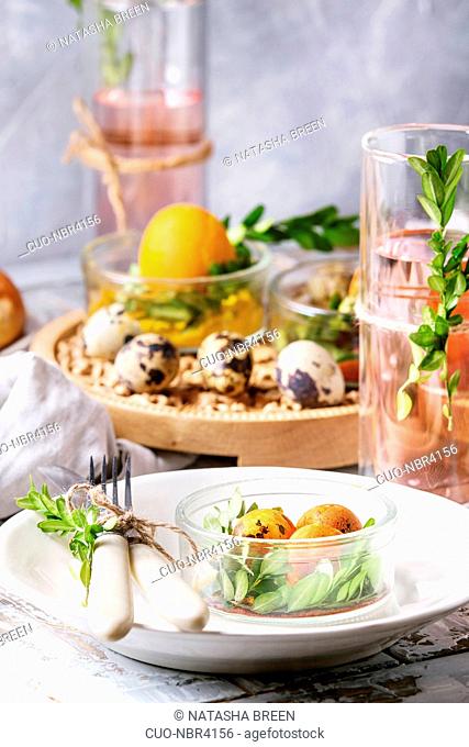 Easter table setting with colored orange eggs, hot cross buns, green branches decorated, empty white plate with cutlery, glass of lemonade drink over white...