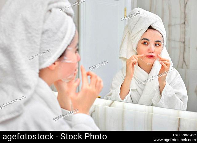 beautiful woman with sheet facial mask in white bathrobes and towel on head in front of mirror in bathroom