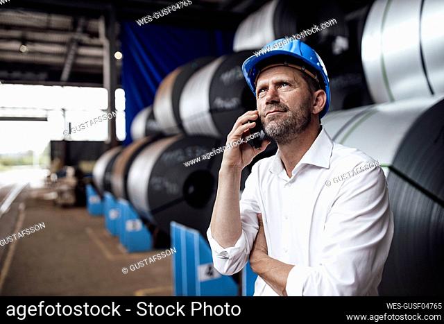 Male owner talking on smart phone looking up while standing against steel rolls in industry