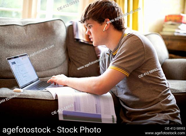 Focused teenage boy with laptop doing homework in living room